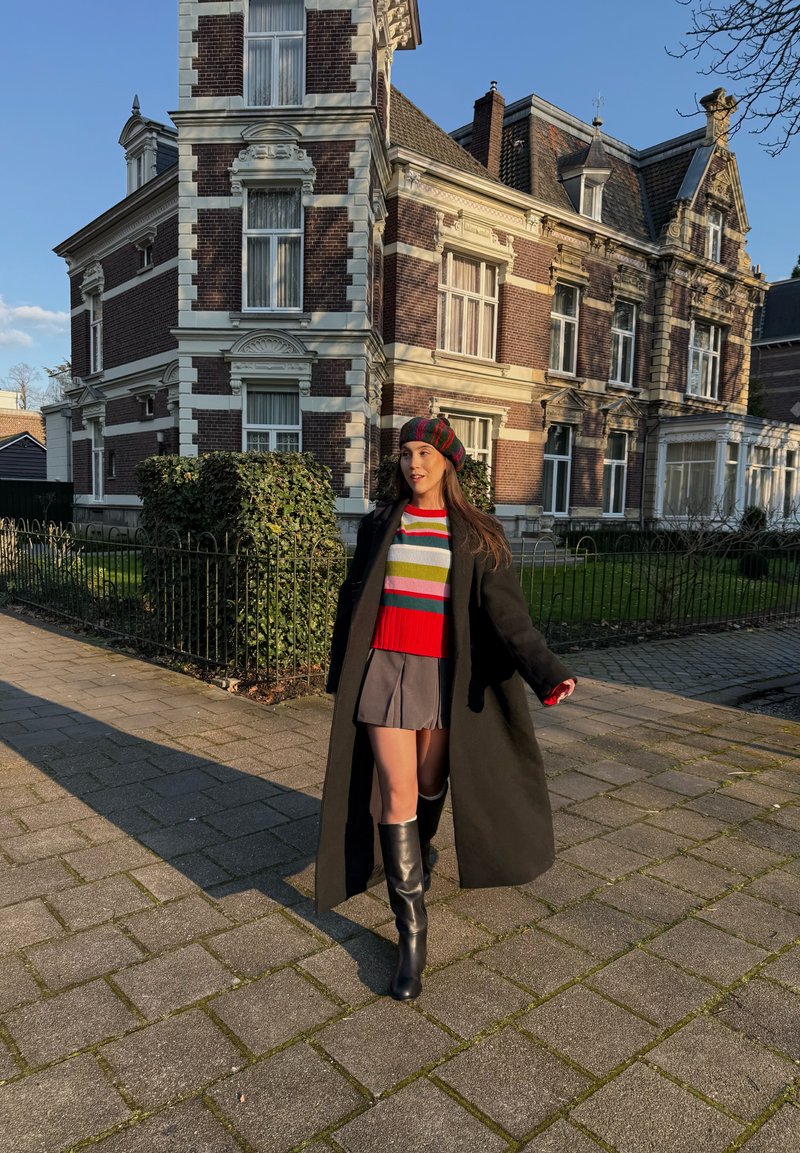 Fekete kabátot, csíkos pulóvert, szürke szoknyát és csizmát viselő nő sétál a járdán egy nagy téglából és kőből épült épület előtt.