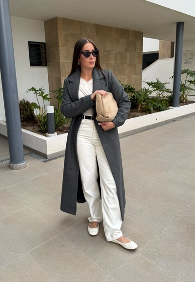 Abrigo gris largo sobre una blusa blanca y pantalones anchos blancos. Sosteniendo un clutch beige. Gafas de sol estilo gato negro y bailarinas blancas.