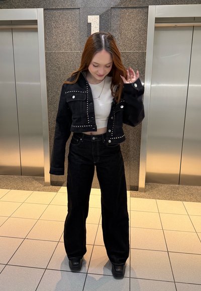 Mujer joven con chaqueta negra tachonada y pantalones de pierna ancha está de pie entre dos ascensores, mirando hacia abajo y tocándose el cabello.