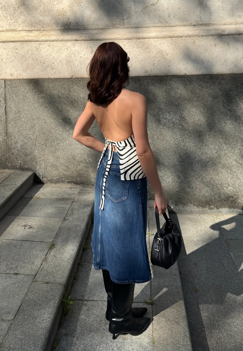 Femme aux cheveux bruns ondulés portant un haut dos nu à imprimé zèbre, une jupe midi en denim, des bottes noires, tenant un sac à main noir, debout sur des marches en pierre.