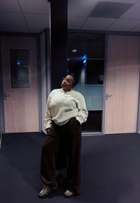 Pullover de creme, calças largas castanhas e ténis cinzentos. Ambiente interior com portas de madeira e iluminação suave. Tapete escuro.