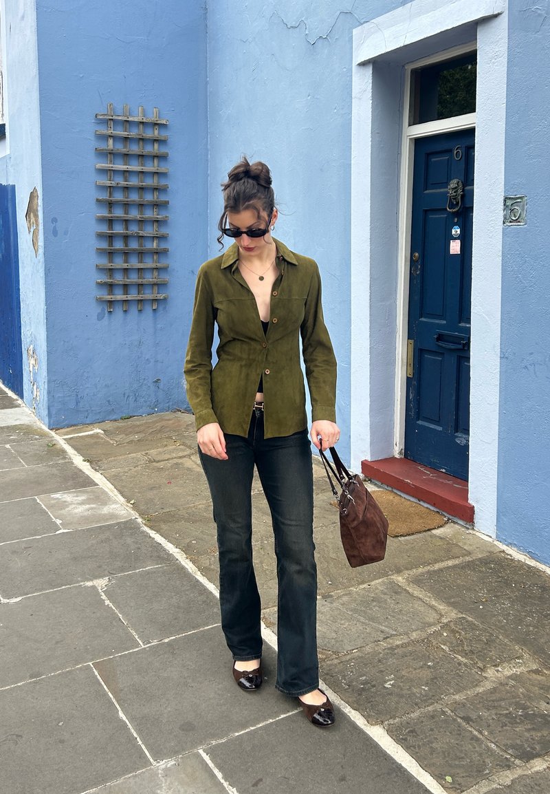 Femme portant des lunettes de soleil, une chemise vert olive, un jean noir et des chaussures marron, marchant sur un trottoir en pierre devant un bâtiment bleu avec une porte bleue.