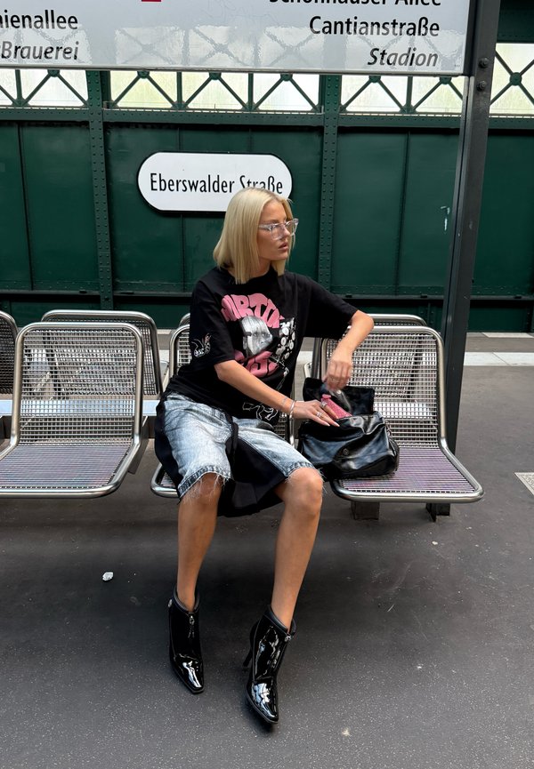 Juodas grafiškas marškinėlis, nusidėvėję džinsiniai šortai, blizgantys juodi kulkšnies batai. Sėdi ant metalinio suolo transporto stotyje. Juoda rankinė rankoje.