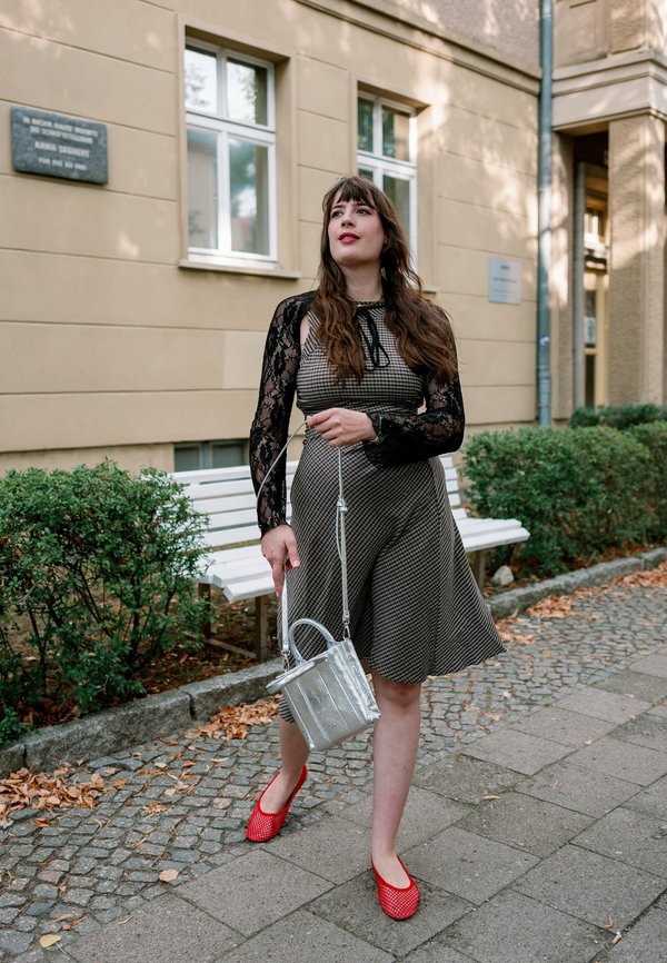 Femme aux longs cheveux bruns portant une robe à motifs et des chaussures rouges tient un sac à main argenté en marchant sur un trottoir près de verdure et de bancs.