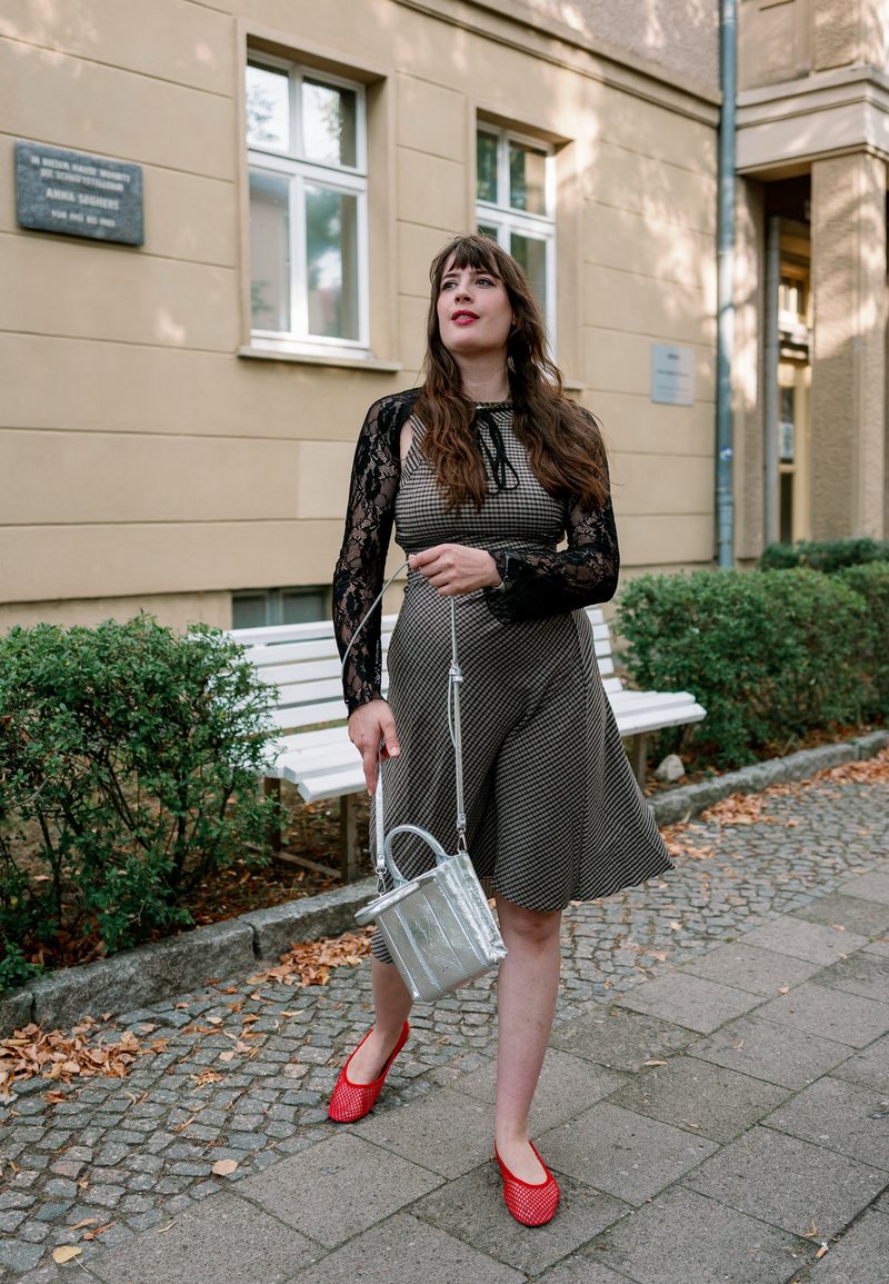 Frau mit langem braunem Haar, die ein gemustertes Kleid und rote Schuhe trägt, hält eine silberne Handtasche, während sie auf einem Bürgersteig in der Nähe von Grünflächen und Bänken geht.