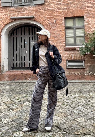 Mujer vistiendo gorra blanca, chaqueta negra, camisa beige, pantalones de cuadros, zapatos blancos, sosteniendo un bolso negro, de pie en una calle de adoquines cerca de un edificio de ladrillo.
