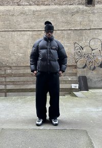 Schwarze Daunenjacke mit hohem Kragen, kombiniert mit weiten schwarzen Hosen. Die Person trägt eine schwarze Mütze und eine Sonnenbrille, dazu graue Sneakers.