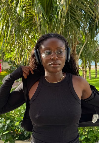 Mujer joven con gafas y auriculares ajusta su chaqueta negra al aire libre, con palmeras y vegetación de fondo.