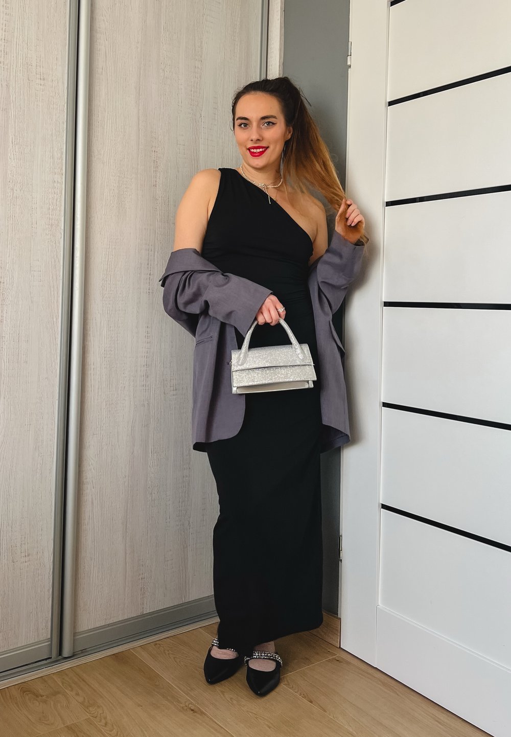 Ένα μαύρο φόρεμα με έναν ώμο σε στρώσεις με μια γκρίζα oversized σακάκι. Κρατά μια μικρή τσάντα από ασήμι με γκλίτερ και φορά μαύρες φλατ πα πούτσια με διακοσμητικά.