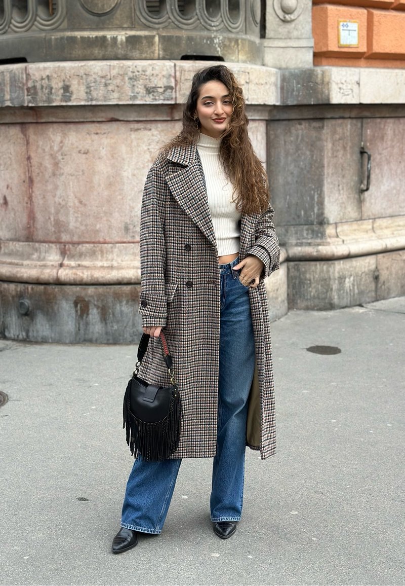 Καρό μακριά παλτό σε μπεζ και καφέ, κοντό λευκό πουλόβερ, φαρδιά μπλε τζιν, μαύρη τσάντα χειρός με φουντάκια και μαύρα μυτερά παπούτσια.