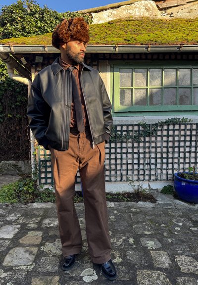 Chaqueta bomber de cuero negra, camisa marrón y pantalones marrones. Gorra de piel en tonos marrones. Zapatos negros. El fondo presenta una ventana verde y una pared de ladrillo.