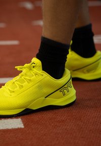 Rumene športne superge z mrežastim zgornjim delom, črnimi poudarki, ravnimi vezalkami in oblazinjenim podplatom, nošene z rdečo stezo in črnimi nogavicami.