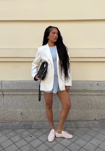 Mujer con el pelo largo trenzado, vistiendo blazer blanco, vestido azul a cuadros, zapatillas rosas, sosteniendo bolso negro, de pie junto a una pared beige.