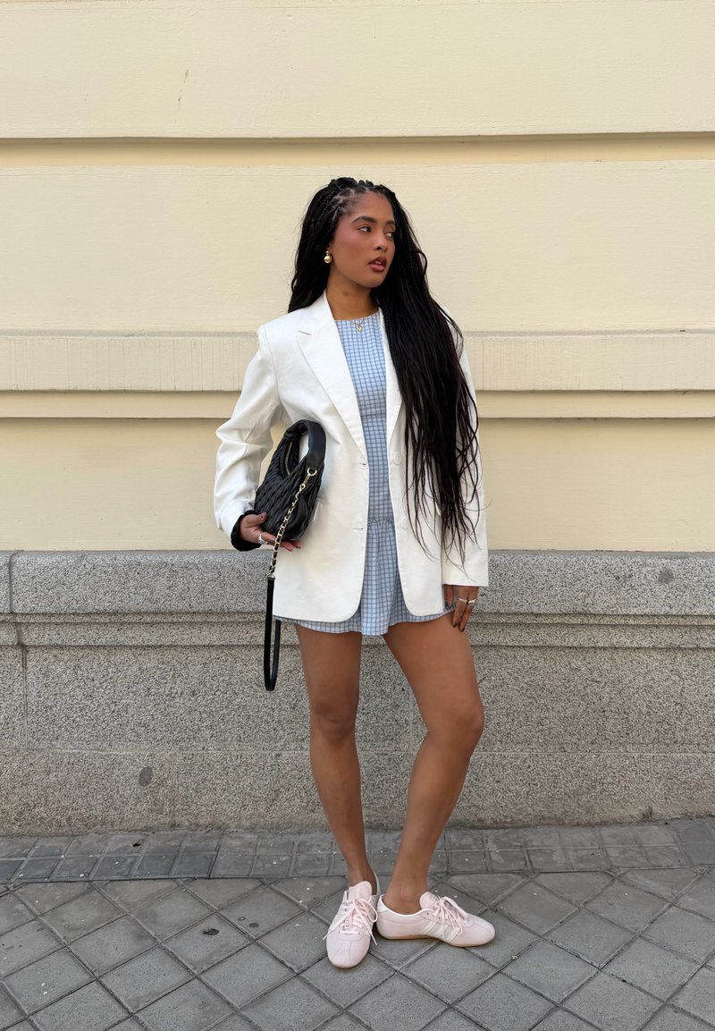 Woman with long braided hair wearing white blazer, blue checkered dress, pink sneakers, holding black handbag, standing by beige wall.