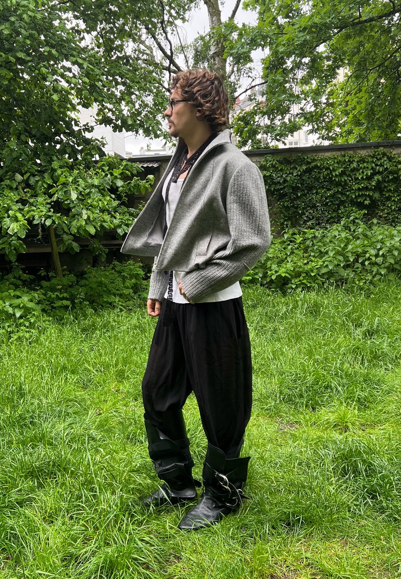 Personne portant une veste grise, une chemise blanche, un pantalon noir ample et de grandes bottes noires, debout sur l'herbe avec des arbres et des buissons verts en arrière-plan.