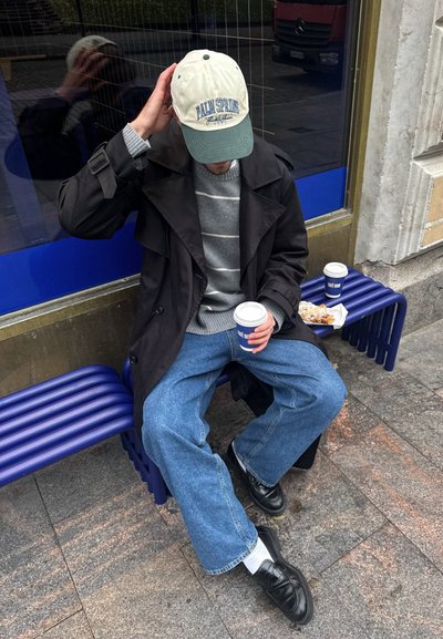 Un abrigo negro sobre un suéter gris a rayas, combinado con unos vaqueros de pernera ancha azul y zapatos negros. Una gorra beige y una taza de café completan el look.