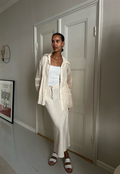 Camisa de lino crema sobre una camiseta blanca, combinada con una falda beige clara y sandalias blancas, con un fondo de pared neutra.