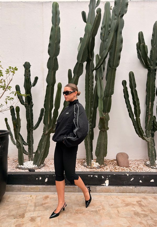 Kvinde iført sort sporty outfit og solbriller, der går forbi høje grønne kaktusser mod en hvid væg som baggrund.