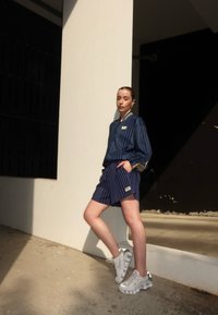 Marine-striped jakke med ribbet krage og matchende shorts; har et logomerke. Båret med sølvfargede joggesko, plassert foran en lys vegg.