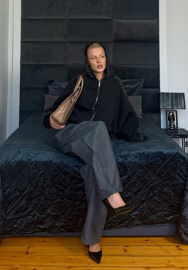 Γυναίκα που φοράει μαύρο φούτερ με κουκούλα και γκρι παντελόνι, καθισμένη σε κρεβάτι με μαύρα σεντόνια, κρατώντας ένα καφέ τσάντα και κοιτάζοντας στο πλάι.