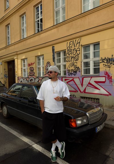 Camiseta blanca de gran tamaño, pantalones cortos negros y zapatillas verdes que lleva un hombre junto a un Mercedes negro, con una pared amarilla cubierta de graffiti al fondo.