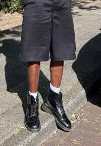 Svarte lær snørebåndstøvler med tykk såle, brukt med hvite ankelstrømper, kombinert med shorts i kne-lengde i svart. Teksturert fortau i bakgrunnen.
