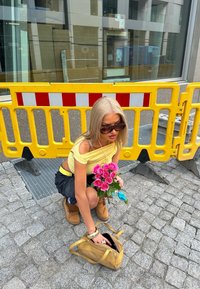 Keltainen kukkaruukku, jossa on vaaleanpunaisia ruusuja, yllään keltainen toppi ja mustat hameet. Kultainen käsilaukku hänen vieressään kivipinnalla, taustalla keltainen este.