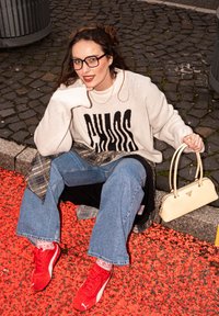 Pull blanc avec l'inscription "CHAOS" en lettres noires, jean bleu ample, baskets rouges et sac à main crème. Sur une surface rouge texturée avec des pierres.