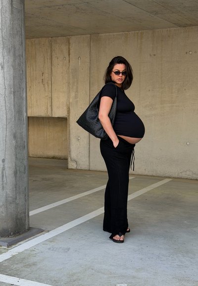 Mujer embarazada con ropa negra y gafas de sol de pie con las manos en los bolsillos, llevando una gran bolsa negra en un aparcamiento de hormigón vacío.