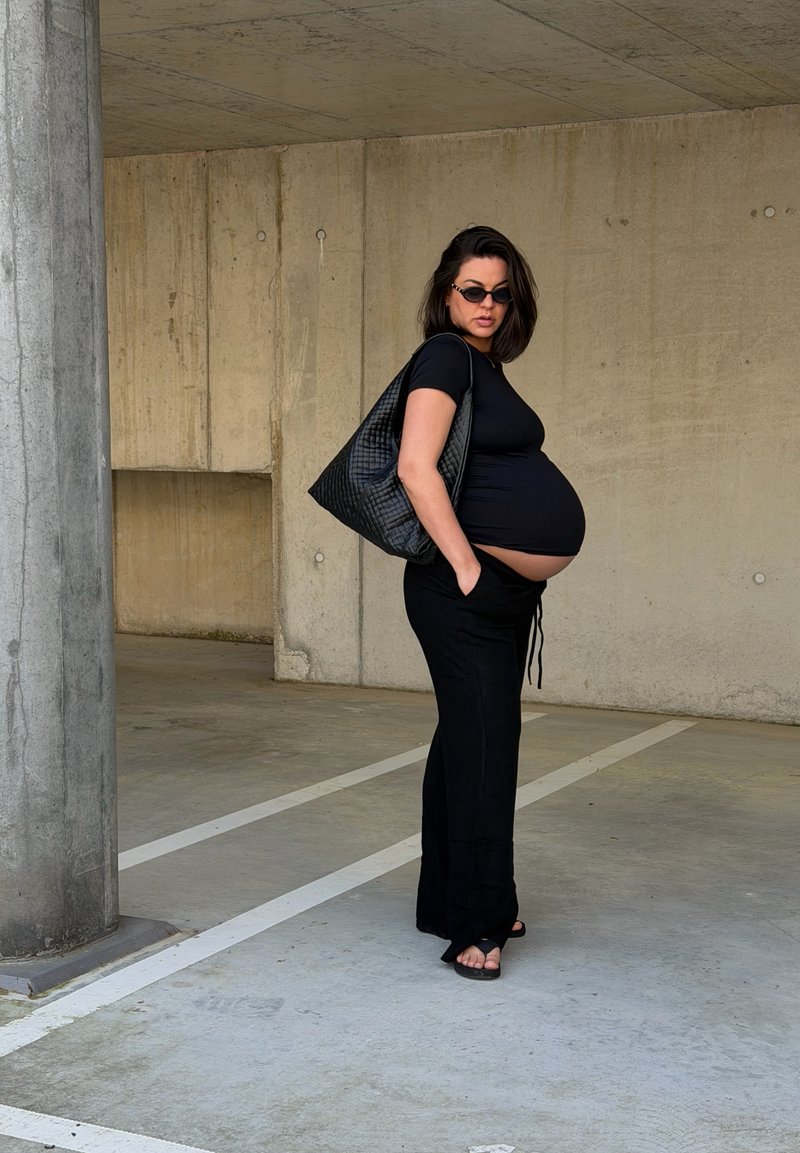 Femme enceinte en tenue noire et lunettes de soleil, debout les mains dans les poches, portant un grand sac noir dans un parking en béton vide.
