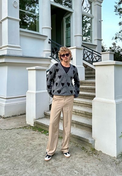 Cárdigan gris con patrones de corazones negros, camiseta negra debajo, pantalones de pierna ancha beige y zapatillas en blanco y negro, de pie en escalones de piedra.