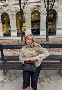 Beige trenssitakki, jossa on korkea kaulus, ja tummat, lyhyet housut. Henkilöllä pieni, tummanruskea käsilaukku. Puistopenkki, jossa on lainaus ranskaksi.