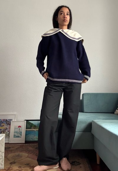 Mujer de pie en el interior con un suéter azul marino con un gran cuello blanco, pantalones anchos negros y zapatillas rosas, cerca de un sofá verde azulado y obras de arte enmarcadas.