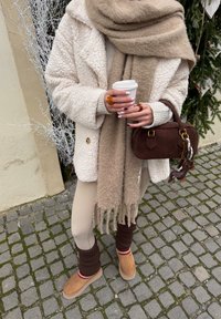 Personne portant un manteau beige, une écharpe et des leggings, tenant un gobelet à emporter et un sac à main marron foncé avec un porte-clés en forme de petit animal.