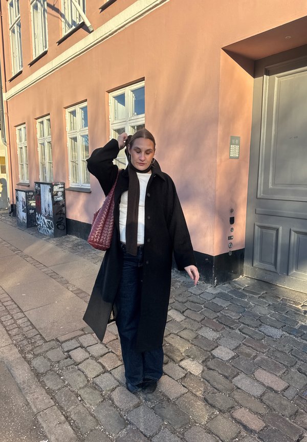 Sieviete ar tumšu garu mēteļu un šalli staigā pa bruģētu ietvi blakus persiku krāsas ēkai ar lieliem logiem un pelēkām durvīm.