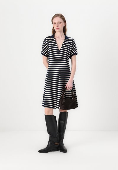 Vestido a rayas con mangas cortas en azul marino y beige, escote en V, falda acampanada; combinado con botas negras hasta la rodilla y un bolso de mano marrón oscuro.