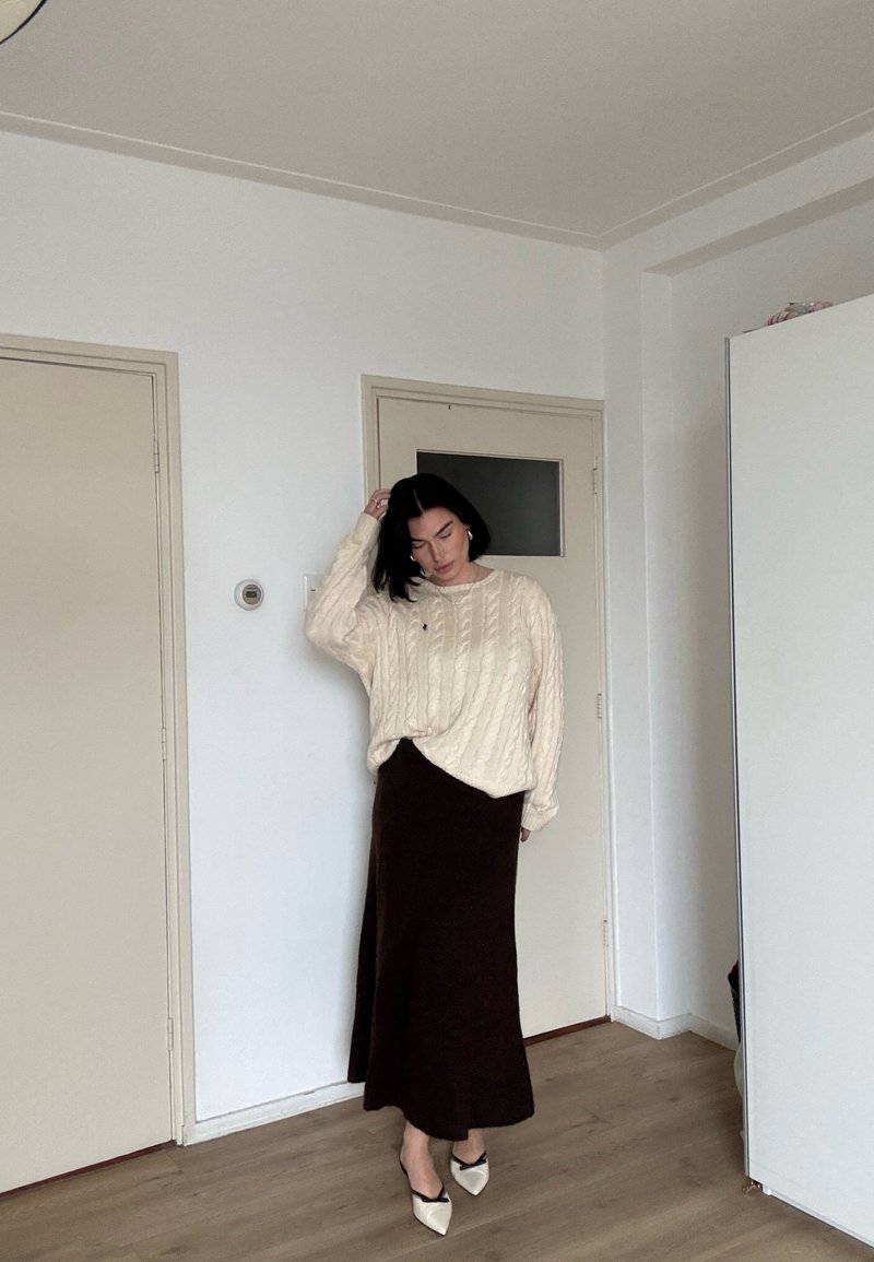 Femme aux cheveux noirs portant un pull crème à torsades, une longue jupe foncée et des chaussures blanches pointues, debout dans une pièce minimaliste avec des portes fermées.