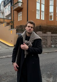Jeune homme portant un manteau noir avec une écharpe grise nou ée autour du cou, debout dans la rue devant un bâtiment brun aux fenêtres blanches.