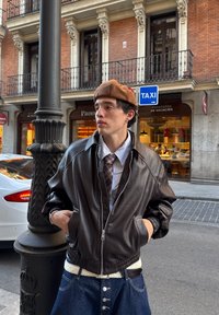 Brązowa skórzana kurtka bomberka z prążkowanymi mankietami i ściągaczem u dołu, noszona na jasnoniebieskiej koszuli w paski i wzorzystym krawacie, w połączeniu z dżinsami.