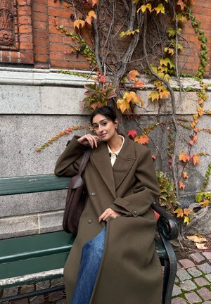 Abrigo de lana marrón con diseño cruzado, colocado sobre un banco verde, con hojas otoñales y un fondo de ladrillos.