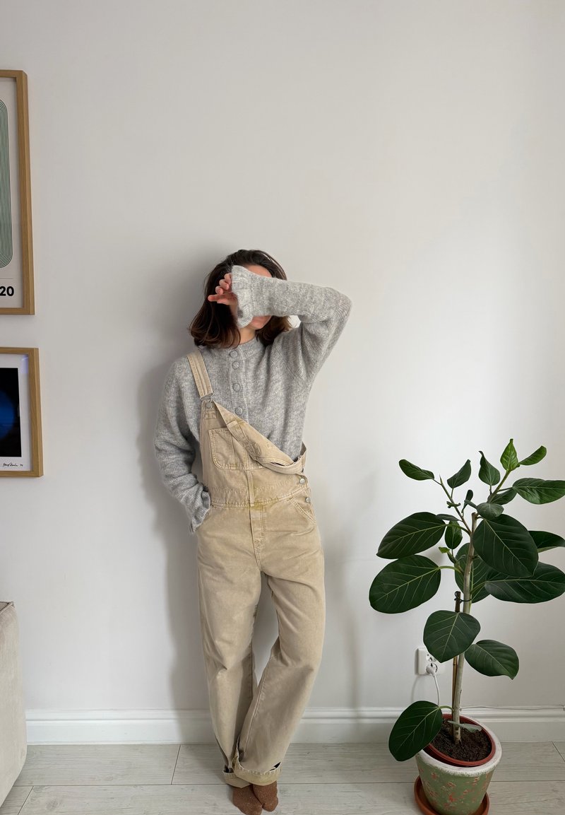 Person klädd i beige arbetsrock och grå tröja, står med en arm som täcker ansiktet, bredvid en bladig krukväxt och inramad väggkonst.