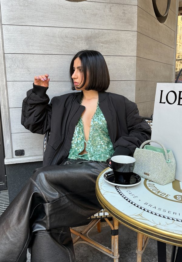 Giubbotto bomber nero, top verde con paillettes e pantaloni di pelle neri. Piccola borsa di perline con manico verde menta su un tavolo di caffè con caffè.