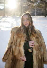 Femme aux longs cheveux ondulés portant un grand manteau en fourrure marron, debout dans une forêt enneigée avec la lumière du soleil filtrant à travers les arbres derrière elle.