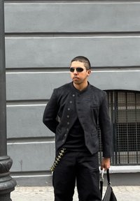 Black textured jacket featuring a high collar and buttoned front, teamed with a black shirt and trousers. Distinctive detail: a chain embellishment on the side.