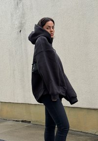 Μαύρος oversized hoodie με κουκούλα και κορδόνι, διαθέτει μπροστινή τσέπη, φορεμένος πάνω από στενές μαύρες κολάν. Φόντο είναι ένας ανοιχτός γκρίζος τοίχος.