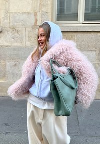 Femme souriante portant une veste rose duveteuse sur un sweat à capuche gris et un pantalon crème, tenant un sac à main vert, debout devant un mur en pierre avec une fenêtre.