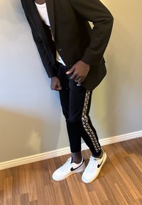 Blazer noir sur un t-shirt blanc, associé à un pantalon de jogging noir avec une bande à motif et des baskets blanches Nike. Sol en bois naturel.