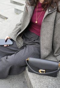 Čierna kožená kabelka so zlatými kovovými detailmi, nosená s šedým tvídovým sakom, bordovým topom a šedými širokými nohavicami. Telefón v ruke.