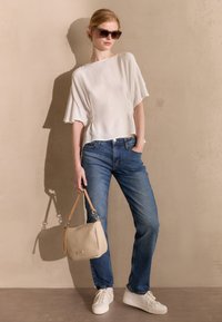 Blusa bianca con maniche ampie, jeans blu e sneakers bianche. Tiene una borsa beige con tracolla. Occhiali da sole scuri completano il look.