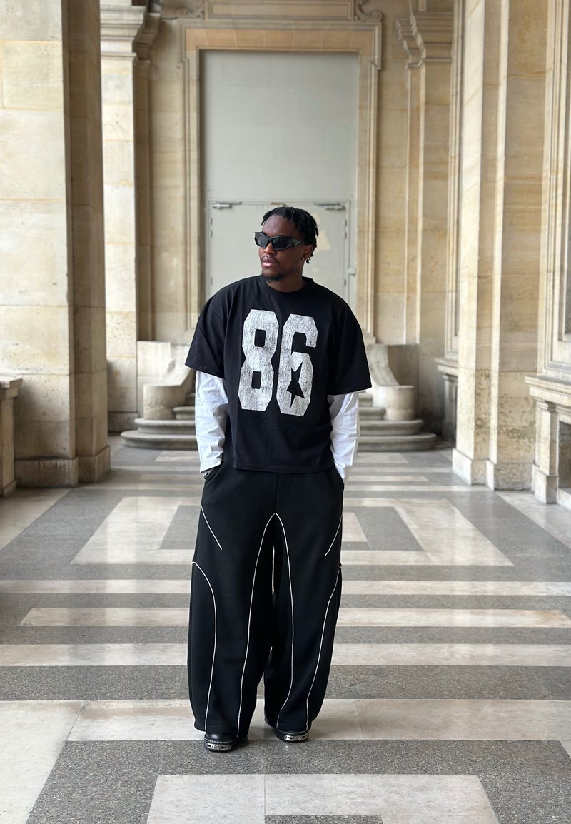 Άνδρας με μαύρα γυαλιά ηλίου, μαύρο μπλουζάκι με τον αριθμό 86, λευκά μακριά μανίκια και φαρδιά μαύρα παντελόνια στέκεται σε μια πέτρινη αίθουσα με γεωμετρικό δάπεδο.